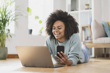 Smiling woman with mobile phone using laptop while lying on floor at home - SBOF03551