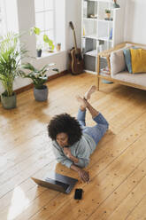 Junge Frau benutzt einen Laptop, während sie auf dem Boden im Wohnzimmer liegt - SBOF03553