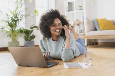 Smiling young woman talking on mobile phone while lying by laptop at home - SBOF03557