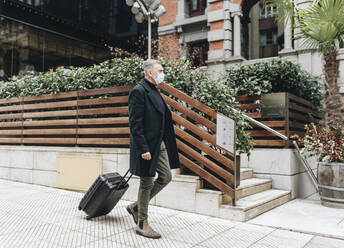 Mature man wearing protective face mask walking with luggage on footpath - DGOF01991