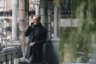 Modischer reifer Mann, der mit seinem Handy telefoniert, während er an einer Glaswand im Hotel steht - DGOF02013