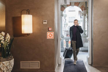 Smiling fashionable man talking on mobile phone while walking with luggage at hotel entrance - DGOF02019