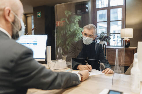 Mann mit Gesichtsschutzmaske beim Ausfüllen eines Formulars an der Hotelrezeption - DGOF02020