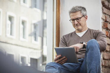 Älterer Mann benutzt ein digitales Tablet, während er zu Hause auf der Fensterbank sitzt - DHEF00620