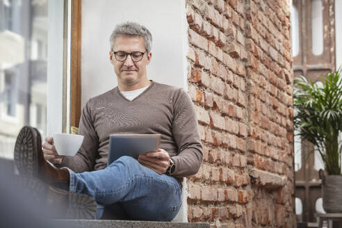 Mann hält Kaffeetasse, während er ein digitales Tablet zu Hause benutzt - DHEF00621