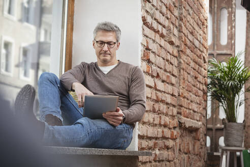 Älterer Mann mit Brille benutzt ein digitales Tablet, während er zu Hause auf der Fensterbank sitzt - DHEF00622