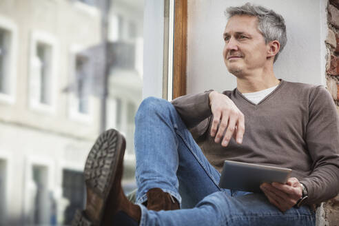 Kaukasischer Mann mit digitalem Tablet, der durch das Fenster schaut, während er zu Hause sitzt - DHEF00624