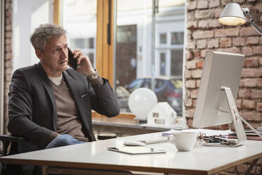 Älterer Geschäftsmann, der am Schreibtisch im Büro sitzt und mit seinem Smartphone spricht - DHEF00625