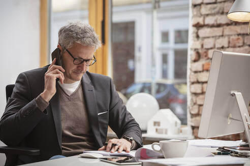 Reifer Geschäftsmann, der ein Dokument betrachtet, während er am Arbeitsplatz mit einem Mobiltelefon spricht - DHEF00628