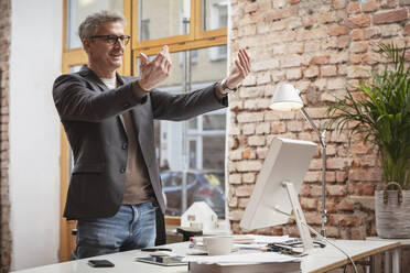 Mature businessman gesturing while standing by desk at work place - DHEF00632