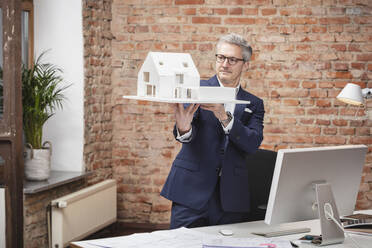 Reifer männlicher Architekt, der ein Modellhaus begutachtet, während er am Schreibtisch im Büro steht - DHEF00635
