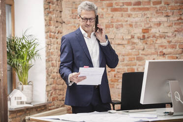 Älterer Geschäftsmann, der mit seinem Smartphone spricht, während er ein Dokument am Schreibtisch im Büro liest - DHEF00640