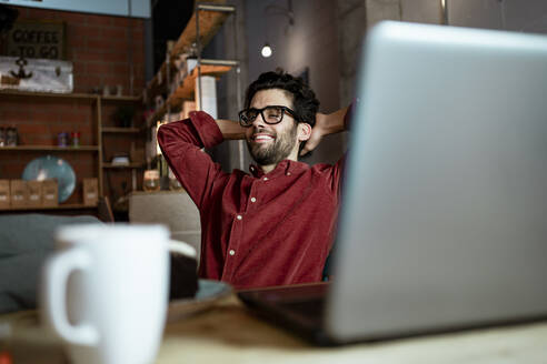 Smiling freelance worker with hands behind head in coffee shop - RCPF00867