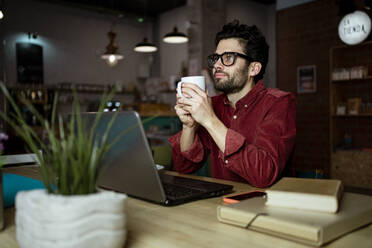 Nachdenklicher männlicher Freiberufler mit Laptop bei einem Kaffee in einem beleuchteten Cafe - RCPF00873