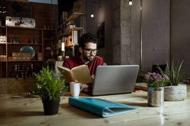 Geschäftsmann arbeitet an einem Laptop und hält ein Buch in einem Cafe - RCPF00876