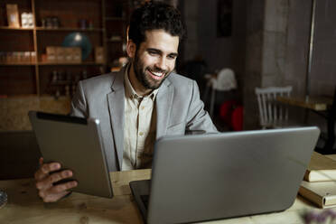 Glücklicher männlicher Freiberufler mit digitalem Tablet bei einem Videogespräch über einen Laptop in einem Café - RCPF00879