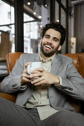 Cheerful businessman holding coffee cup while sitting in coffee shop - RCPF00886