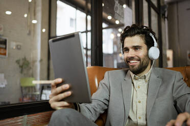 Smiling male entrepreneur listening through headphones while on video call in cafe - RCPF00888