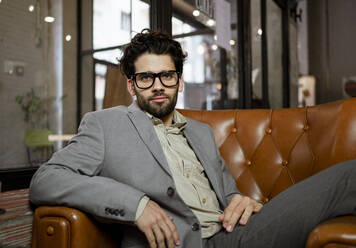 Confident businessman in eyeglasses sitting on arm chair in cafe - RCPF00890