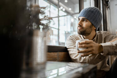 Mittlerer erwachsener Mann mit Strickmütze und Kaffeetasse, der durch das Fenster eines Cafés schaut - RCPF00893