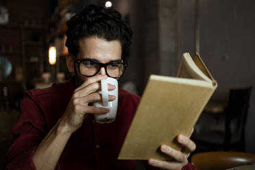 Männlicher Berufstätiger trinkt Kaffee und liest ein Buch in einem beleuchteten Cafe - RCPF00897