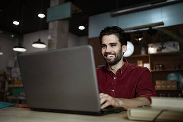 Glücklicher männlicher Unternehmer bei einem Videogespräch über einen Laptop in einem beleuchteten Kaffeehaus - RCPF00900