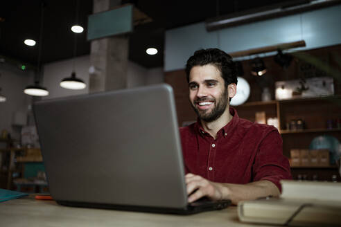 Glücklicher männlicher Unternehmer bei einem Videogespräch über einen Laptop in einem beleuchteten Kaffeehaus - RCPF00900