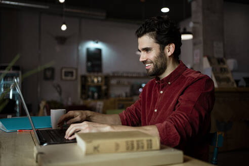 Lächelnder männlicher Unternehmer arbeitet an einem Laptop in einem beleuchteten Café - RCPF00905