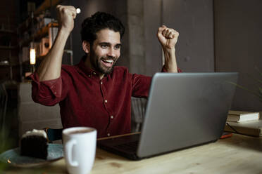 Businessman cheering while looking at laptop while sitting in illuminated coffee shop - RCPF00906