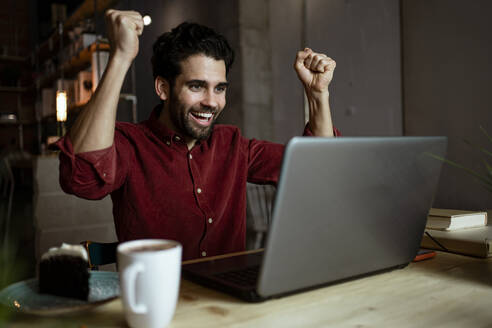 Businessman cheering while looking at laptop while sitting in illuminated coffee shop - RCPF00906