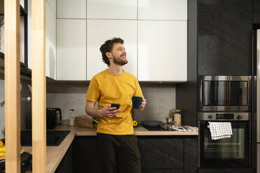 Glücklicher Mann mit Kaffeetasse und Smartphone in der heimischen Küche - VPIF03848