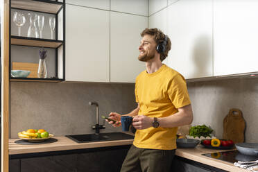 Lächelnder Mann hält Kaffeetasse und Smartphone, während er in der Küche über Kopfhörer Musik hört - VPIF03850