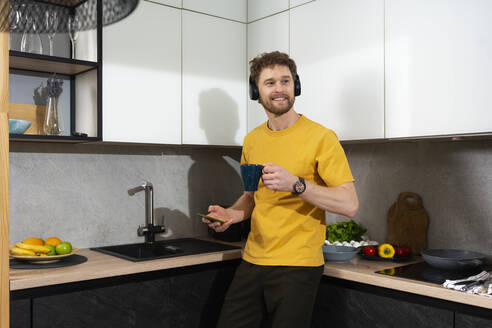 Mann hört Musik und hält Handy und Kaffeetasse in der Küche zu Hause - VPIF03851