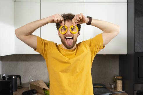Verspielter Mann streckt die Zunge heraus, während er in der Küche Paprikascheiben auf die Augen hält - VPIF03857