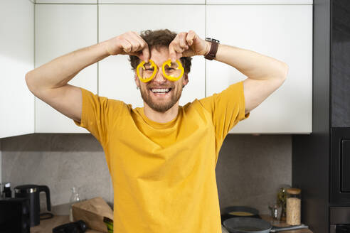 Lächelnder Mann mit Paprikascheiben auf den Augen in der Küche zu Hause - VPIF03858