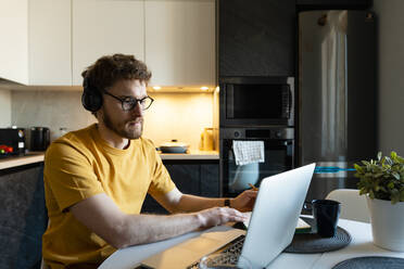 Male freelance worker listening through headphones while using laptop in kitchen - VPIF03862