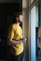 Nachdenklicher Mann, der eine Kaffeetasse hält und durch ein Fenster im Wohnzimmer schaut - VPIF03864
