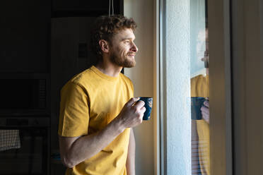 Smiling man looking through window while having coffee at home - VPIF03865