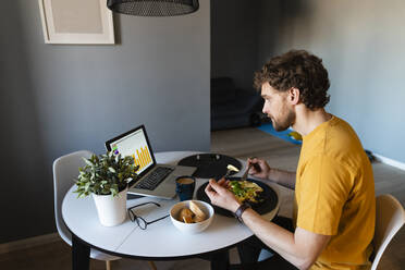 Männlicher Freiberufler beim Essen, während er zu Hause am Laptop arbeitet - VPIF03868
