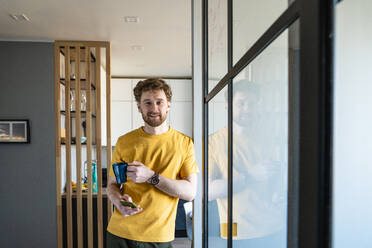 Gut aussehender Mann hält Kaffeetasse und Smartphone, während er sich zu Hause an ein Glas lehnt - VPIF03872