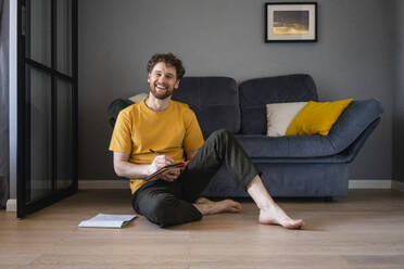 Smiling man sitting with book on floor at home - VPIF03876