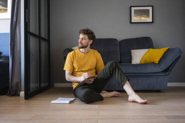 Thoughtful man holding book while sitting on floor in living room - VPIF03877