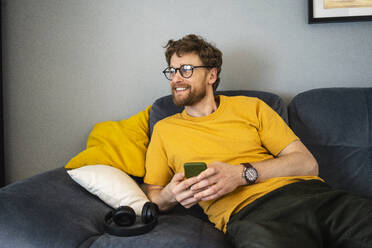 Lächelnder mittelgroßer erwachsener Mann mit Brille und Mobiltelefon, der auf dem Sofa sitzt und wegschaut - VPIF03881