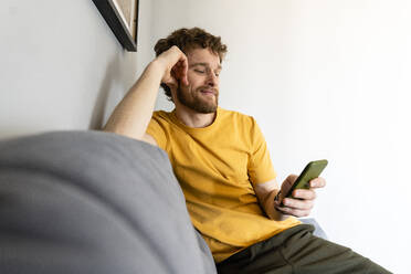 Lächelnder Mann, der ein Mobiltelefon benutzt, während er zu Hause auf dem Sofa sitzt - VPIF03903