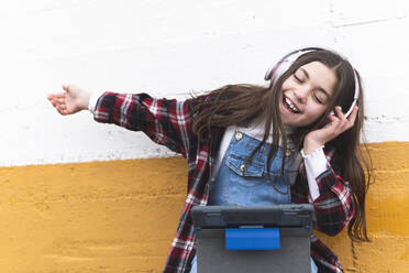 Fröhliches Mädchen hört Musik über Kopfhörer vor einer Wand - JAQF00419