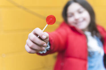 Girl holding red lollipop in front of yellow wall - JAQF00421