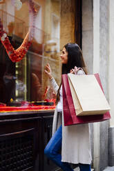 Happy woman with shopping bags looking through glass while window shopping - PGF00478
