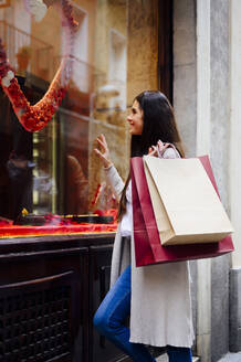Glückliche Frau mit Einkaufstüten, die beim Schaufensterbummel durch Glas schaut - PGF00478