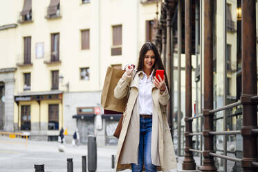 Woman looking at mobile phone holding shopping bags in city - PGF00491