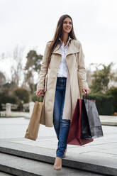 Woman in overcoat moving down from stairs while holding shopping bags - PGF00496